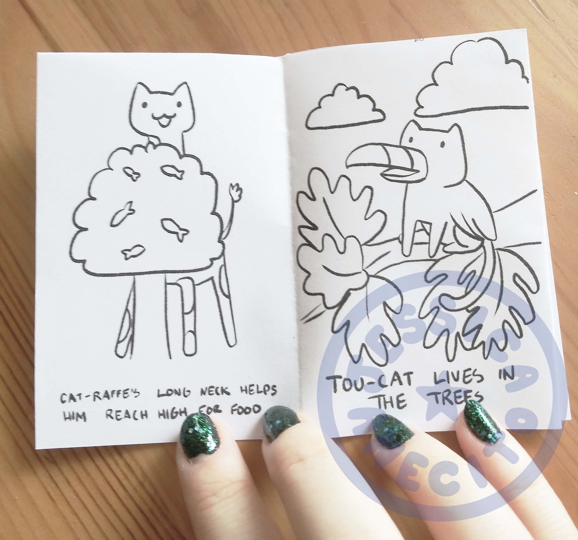 a mini zine is opened to show a picture on the left of the cat-raffe eating from a tree with small fish on the branches. The right page is a picture of a cat with wings and a beak sitting in a tree