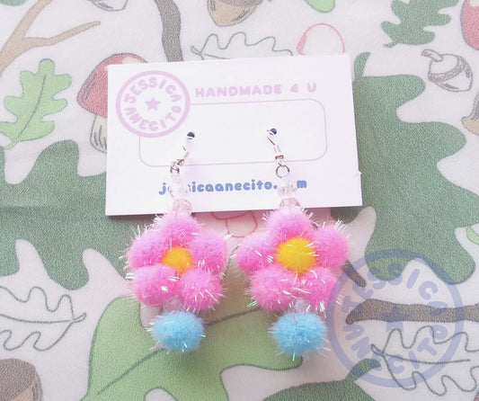 Two earrings are attached to a thick paper that reads, "Handmade 4 u" and "jessicaanecito.com". A logo is on the corner that is shaped like a circle and has the name "Jessica Anecito" going around the inside with a star in the center. The earrings are made of a yellow pom pom surrounded by five pink pom poms to make a flower shape. The flower has a clear bead and a blue pom pom hanging off the bottom and two clear beads on top connecting it to an earring hook.