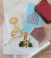 Image is a flat lay of several objects: a red marker, ink pad, a blue notecard that has been stamped with a stamp that says "thank you!", the stamp, a mini booklet, a sticker and some string. The booklet reads, "Catraffe's fall favorites" and shows a cat's head inside a pumpkin on a giraffe body. The sticker is the cat-raffe standing in some leaves and smiling. 